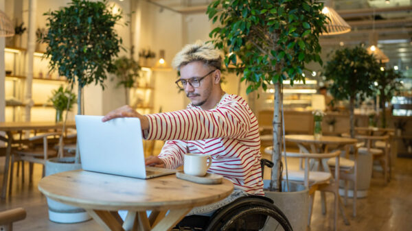 ânăr zâmbitor, cu dizabilități (utilizator de scaun cu rotile manual), lucrând la un laptop într-o cafenea modernă și luminoasă, decorată cu plante mari în ghiveci.