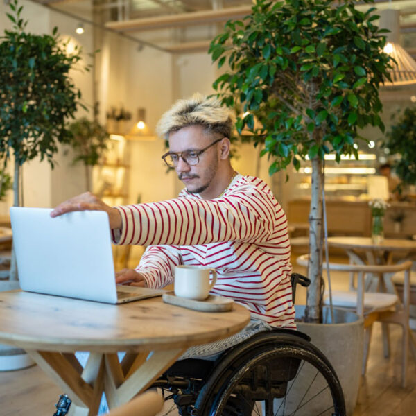 ânăr zâmbitor, cu dizabilități (utilizator de scaun cu rotile manual), lucrând la un laptop într-o cafenea modernă și luminoasă, decorată cu plante mari în ghiveci.