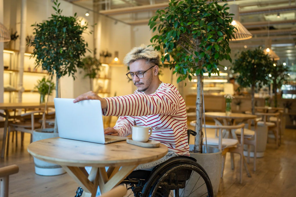 ânăr zâmbitor, cu dizabilități (utilizator de scaun cu rotile manual), lucrând la un laptop într-o cafenea modernă și luminoasă, decorată cu plante mari în ghiveci.